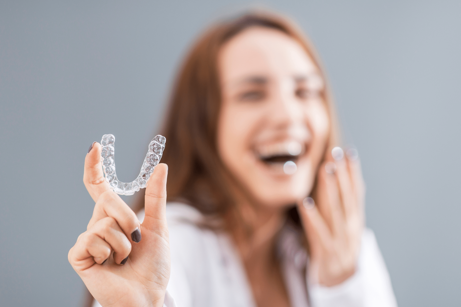 Hand holding a clear aligner tray with a blurry smiling woman in the background