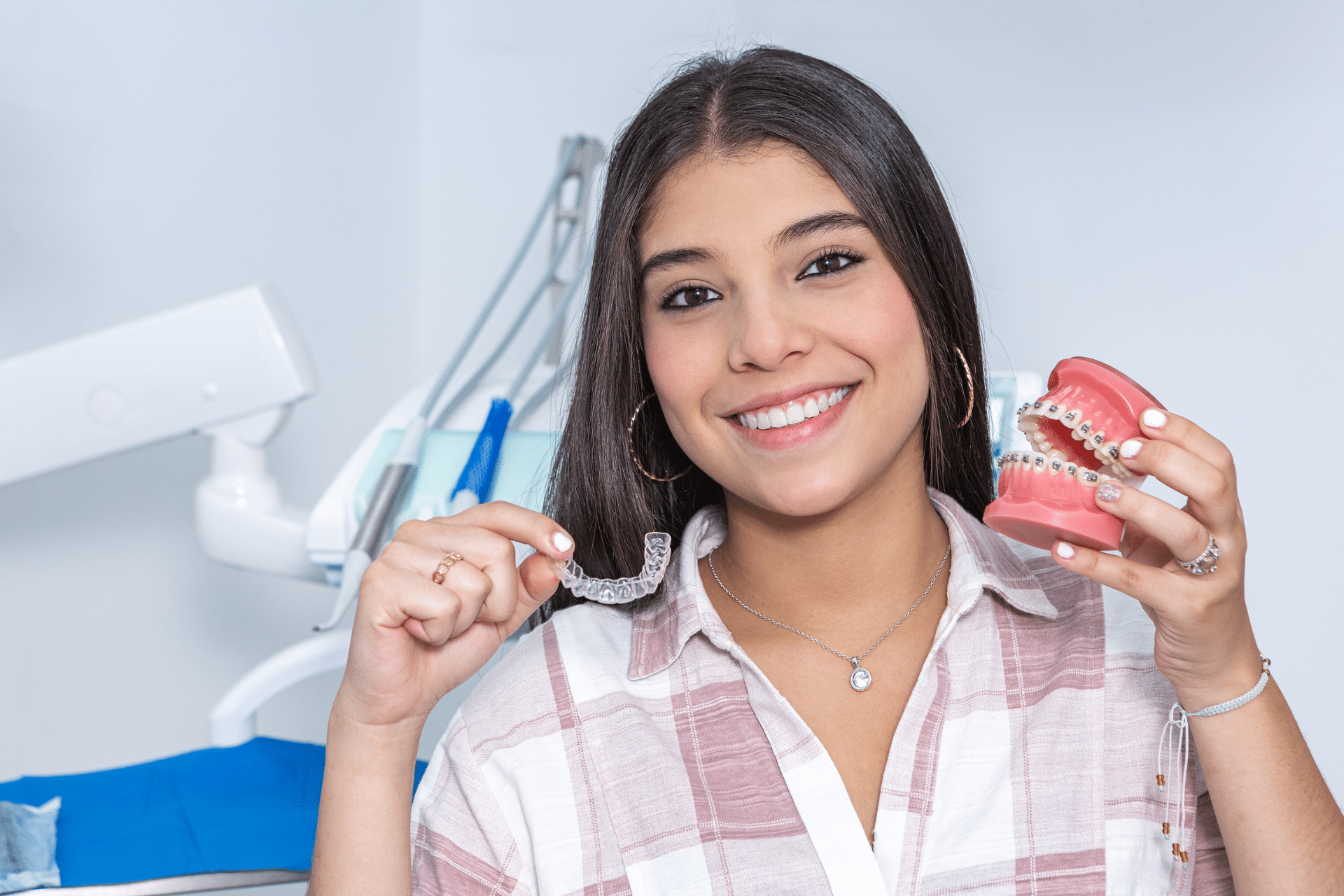 Smiling woman holding a clear dental aligner and a model of teeth with metal braces.